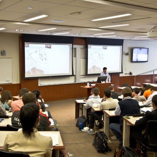 A lecture hall with a person presenting at the front, using a computer and dual projection screens displaying diagrams. Students are seated, attentively taking notes.
